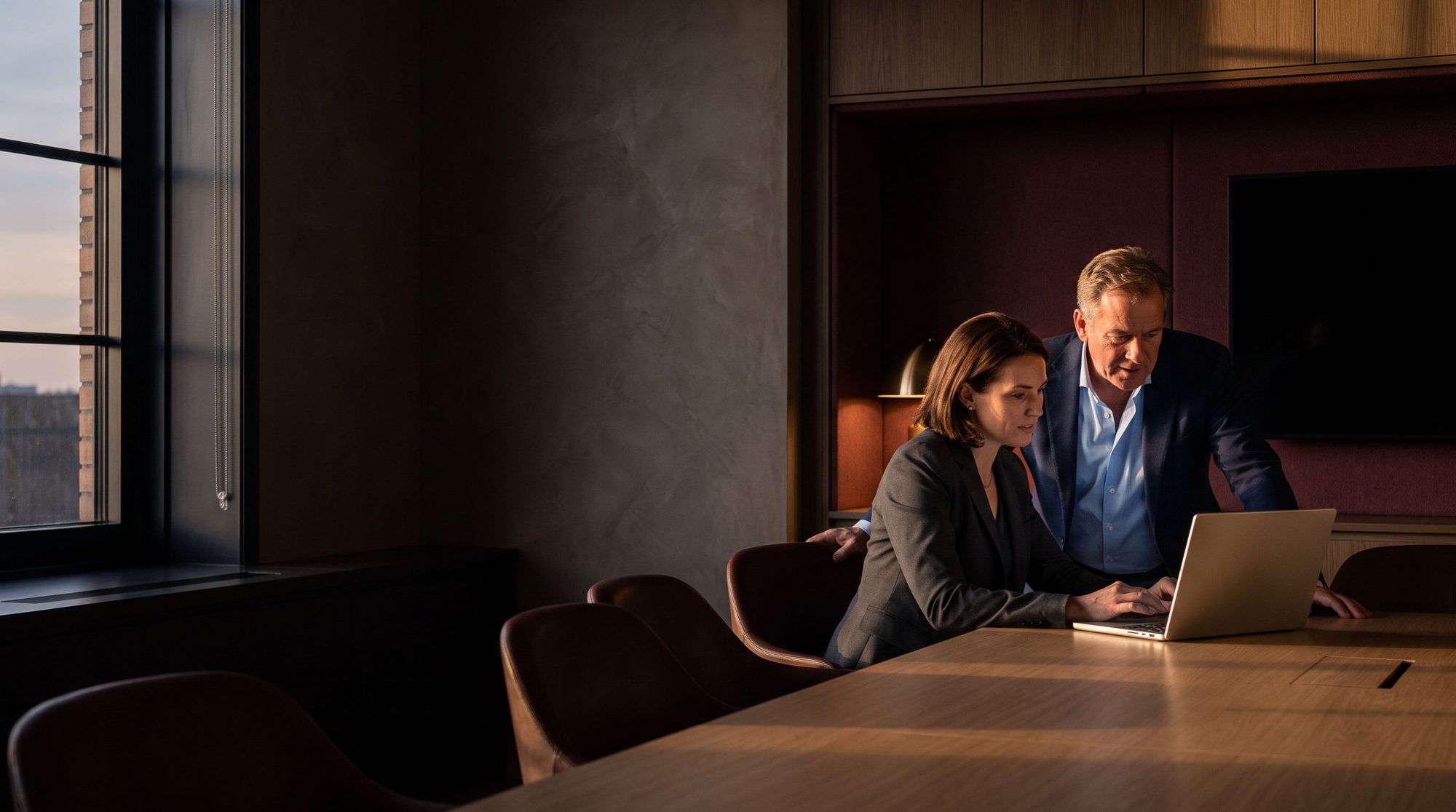 A business owner and a tax advisor leaning in over a laptop in a warm, modern meeting room at dusk.